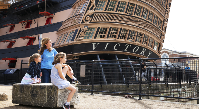 A mother and two children sit outside HMS Victory, a wooden ship. 