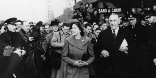 A young Queen Elizabeth visiting a Naval base surrounded by Naval officers and press. 