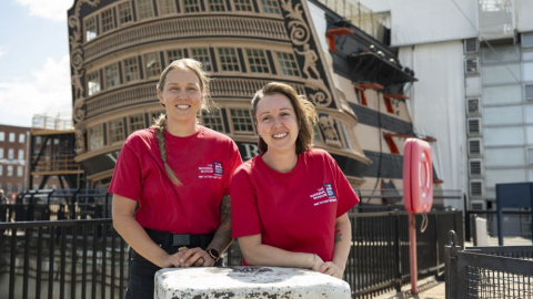 Two female shipwrights stand next to HMS Victory's stern.