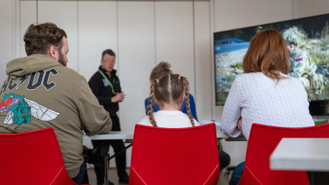 Families seated on red chairs watching a museum educator and a screen showing a camouflaged Royal Marine