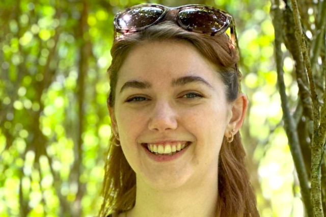 A young white woman with brown hair standing in a forest. 