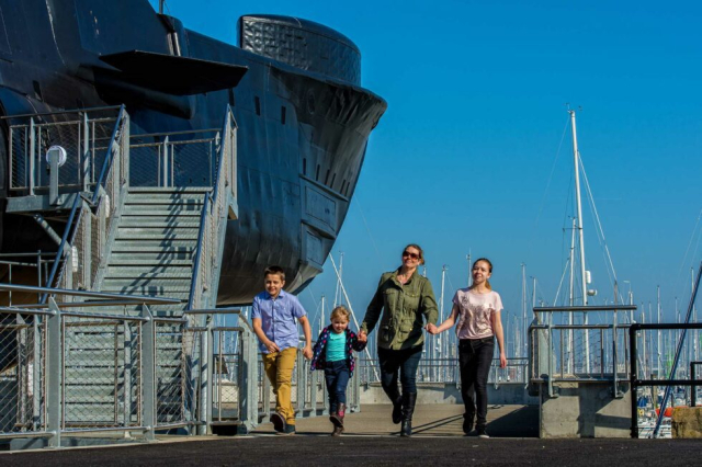 A mother with her children stroll hand in hand next to HMS Alliance. 