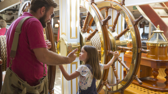 A father and daughter standing next to HMS Warrior's interior wheel.