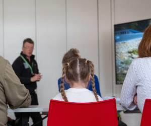 Families seated on red chairs watching a museum educator and a screen showing a camouflaged Royal Marine